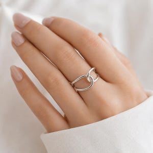 May include: A polished silver ring featuring a bypass design with two smooth, interlocking bands that cross over each other at the center. The ring has a high-shine metallic finish and a continuous, fluid shape that wraps around the finger.
