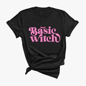 May include: A black short-sleeved t-shirt featuring the text "Basic witch" in a pink and white checkered pattern. The lettering is in a bold, rounded font, and a small pink bat icon is positioned above the word "Basic" on the front of the shirt.