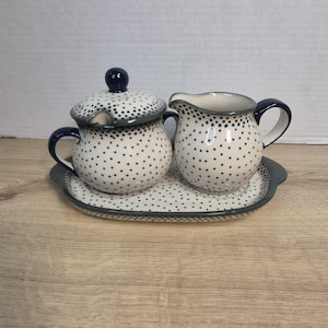 Może przedstawiać: Ceramiczna cukiernica z pokrywką, dzbanek na śmietankę oraz owalna taca do serwowania. Każdy element jest w kolorze kremowym z wzorem w małe, ciemnoniebieskie kropki oraz ciemnoniebieskimi obrzeżami. Cukiernica posiada dwa uchwyty i gałkę na pokrywce.