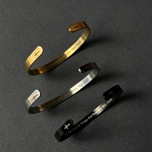May include: Three open cuff bracelets in gold, silver, and black tones. Each bracelet features a small cross symbol at the end and the engraved text "With God All Things Are Possible Mat 19:26" along the inner band of the metal cuff jewelry.