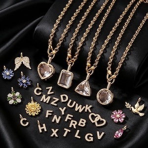 May include: A collection of gold-tone jewelry including four necklaces with heart, rectangular, triangular, and circular clear pendants, alongside various charms shaped as letters, flowers, wings, a bird, and hearts, all featuring small clear stones.