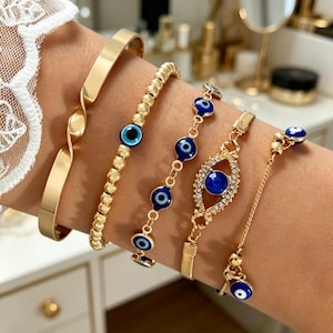 Könnte beinhalten: Ein Set aus fünf goldfarbenen Armbändern. Eines ist ein glatter, gedrehter Armreif, eines eine Perlenkette, zwei zeigen blau-weiße Nazar-Amulette und eines enthält ein zentrales Nazar-Amulett, umgeben von kleinen klaren Kristallen an einer Kette.