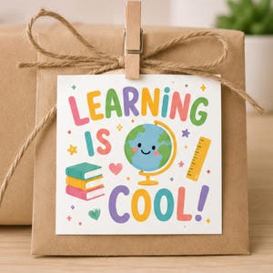 Peut inclure: Une étiquette cadeau carrée blanche fixée sur un paquet en papier kraft avec une pince en bois et de la ficelle. L'étiquette présente le texte coloré LEARNING IS COOL! avec des illustrations de trois livres, un globe souriant et une règle jaune.