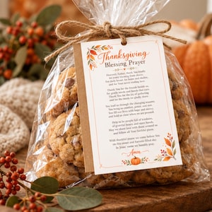 May include: A clear plastic bag filled with chocolate chip cookies, tied with a brown twine bow. A rectangular kraft paper tag is attached, featuring the text Thanksgiving Blessing Prayer, a printed poem, and colorful autumn leaf and pumpkin graphics.