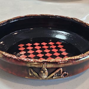 May include: A round, shallow bowl with a glossy black interior featuring a red, four-petaled floral pattern at the center. The exterior is a mottled reddish-brown with a textured, gold-toned rim and a central gold-toned emblem featuring a key.