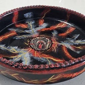 May include: A round black ceramic serving dish featuring a central circular emblem with a red and black face design. The interior surface is decorated with radiating yellow and white lightning-like patterns, and the rim has a textured red border.