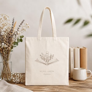 Può includere: Una borsa tote in tela color crema con un'illustrazione a linee marrone scuro raffigurante un libro aperto da cui sbocciano fiori di campo. Sotto l'illustrazione, il testo recita: READ. GROW. dream. La borsa è dotata di due lunghi manici.