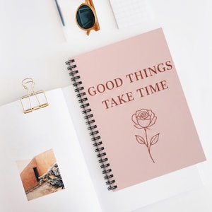 Puede incluir: Un cuaderno de espiral con una cubierta de color rosa claro que presenta el texto en rojo oscuro GOOD THINGS TAKE TIME y una ilustración de una rosa. El cuaderno está abierto en una página que muestra una pequeña foto cuadrada de un edificio y un clip dorado.