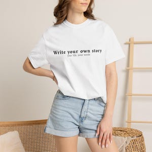 Può includere: Una t-shirt bianca a maniche corte con il testo stampato in nero "Write your own story" sul petto, con il sottotitolo più piccolo "your life, your words" direttamente sotto la frase principale, abbinata a pantaloncini di jeans azzurri.
