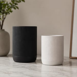 May include: Two cylindrical containers, one black and one white, featuring a vertical ribbed texture on the upper portion and a smooth, solid base. The containers are open at the top and have a uniform, straight-sided shape with rounded bottom edges.
