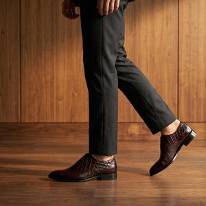 Può includere: Un paio di scarpe eleganti in pelle marrone scuro con una trama testurizzata effetto pelle di coccodrillo, dotate di tacco basso, punta arrotondata e lacci sottili scuri, indossate con pantaloni neri attillati che terminano sopra la caviglia.