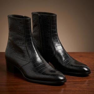 Könnte beinhalten: Ein Paar schwarze Stiefeletten mit einer strukturierten Lederprägung in Krokodiloptik. Diese Stiefel haben eine schlanke Silhouette, eine spitz zulaufende Zehenpartie, einen niedrigen Blockabsatz und einen vertikalen Reißverschluss an der Innenseite.