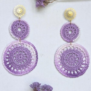 May include: A pair of dangle earrings featuring gold-tone filigree studs connected to two circular, crocheted lavender and white ombre motifs. The design consists of a smaller top circle and a larger bottom circle, both with intricate stitching.