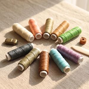 May include: A collection of eleven cylindrical spools of waxed thread in various colors including cream, peach, olive, tan, green, charcoal, sage, brown, turquoise, and purple, arranged in a semi-circle with a small metal tool and a bone folder.