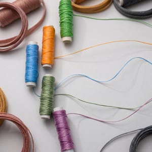 May include: A collection of waxed thread spools in vibrant colors including green, orange, blue, olive, and purple, accompanied by coiled lengths of matching cord. Each spool features a central core with thread wound tightly around the exterior.