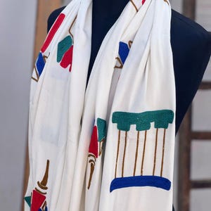 Może przedstawiać: Biały szal z haftowanymi geometrycznymi wzorami w kolorze czerwonym, niebieskim, zielonym i brązowym. Projekt zawiera powtarzające się motywy stylizowanych struktur i kształtów rozmieszczonych na tkaninie, tworząc teksturowany i kolorowy wygląd.