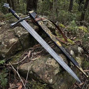 May include: A double-edged sword with a patterned Damascus steel blade, a black hilt, and a crossguard. It is accompanied by a black leather scabbard featuring intricate gold-colored filigree designs and a brown leather strap for carrying.