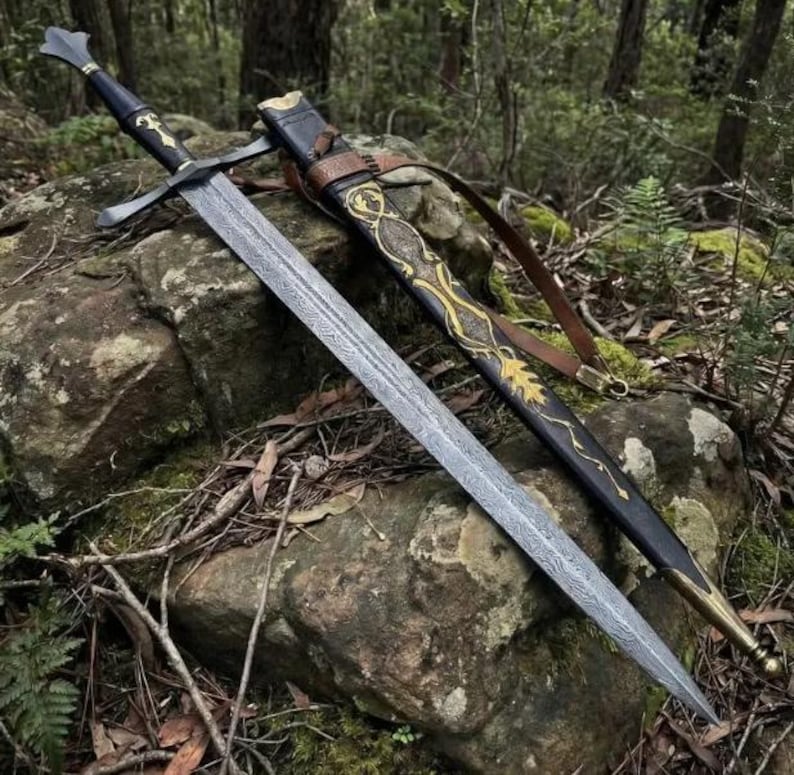 May include: A double-edged sword with a patterned Damascus steel blade, a black hilt, and a crossguard. It is accompanied by a black leather scabbard featuring intricate gold-colored filigree designs and a brown leather strap for carrying.