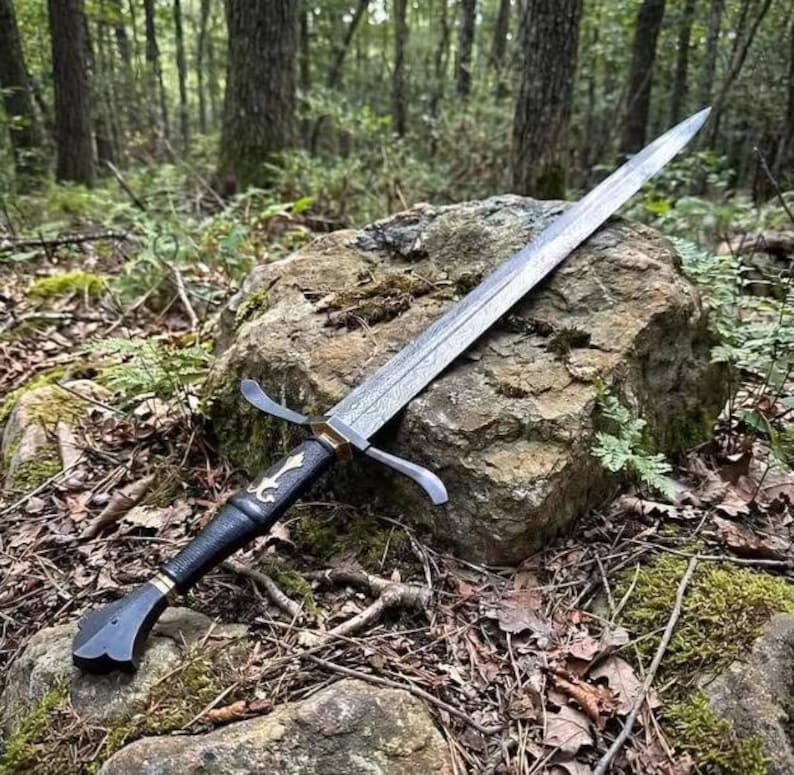 May include: A double-edged steel sword with a textured, dark metal blade featuring a visible pattern. The hilt has a black, textured grip, a silver-colored metal crossguard, and a decorative silver-colored inlay on the handle near the guard.