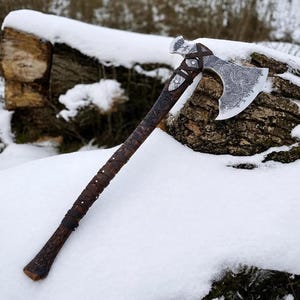 May include: A hand axe with a dark brown wooden handle featuring intricate carvings and leather wrapping. The metal axe head displays detailed etched patterns and symbols, secured to the handle with leather straps and metal rivets.