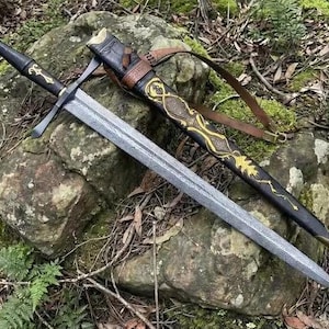 Può includere: Una spada in acciaio a doppio taglio con lama testurizzata, impugnatura rivestita in pelle nera con emblema dorato e fodero nero decorato con motivi in filigrana d'oro, cinturino in pelle marrone e punta in ottone, appoggiata su una roccia.
