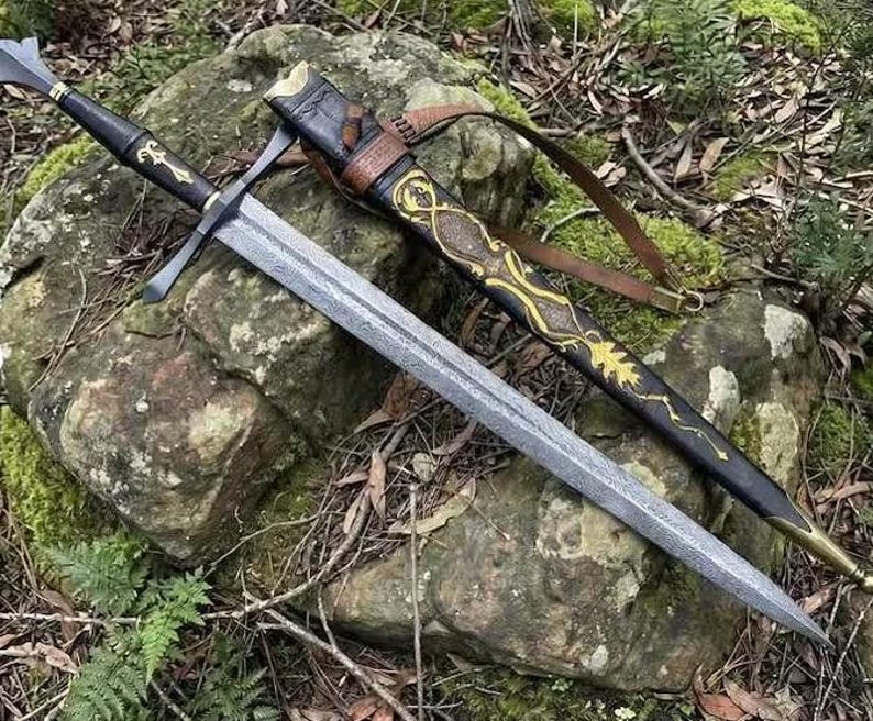 May include: A double-edged steel sword with a textured blade, a black leather-wrapped hilt featuring a gold emblem, and a black scabbard decorated with gold filigree patterns, a brown leather strap, and a brass tip, resting on a mossy rock.