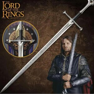 May include: A silver metal sword with a long, pointed blade and a cross-shaped hilt featuring a black grip. The sword is displayed alongside a circular emblem showing a detailed hilt design with gold wings and the text The Lord of the Rings.