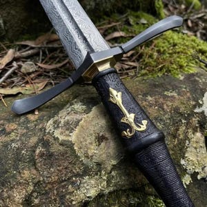 May include: A dagger with a Damascus steel blade featuring a wavy pattern. The hilt has a black textured grip, a brass crossguard, and a central brass fleur-de-lis emblem. The pommel and guard are dark metal, contrasting with the gold-toned accents.