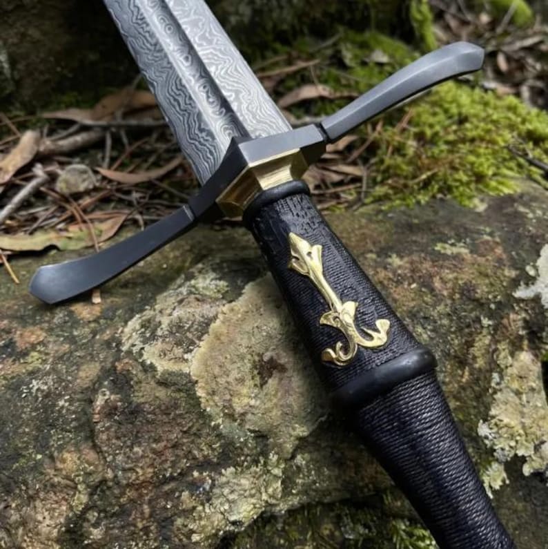 May include: A dagger with a Damascus steel blade featuring a wavy pattern. The hilt has a black textured grip, a brass crossguard, and a central brass fleur-de-lis emblem. The pommel and guard are dark metal, contrasting with the gold-toned accents.