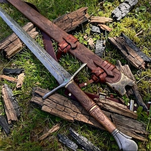 May include: A double-edged sword with a patterned steel blade, a crossguard featuring a small green stone, and a brown leather-wrapped handle. The sword is accompanied by a matching brown leather scabbard with intricate embossed designs.