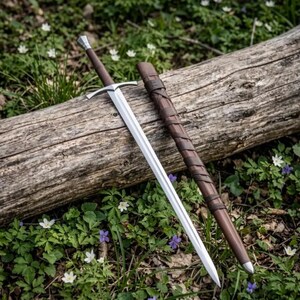 Op de afbeelding: Een tweesnijdend stalen zwaard met een zilverkleurige pareerstang en een bruin met leer omwikkeld handvat, vergezeld van een bijpassende bruine lederen schede met decoratieve leren veters die om de buitenkant van de schede zijn gewikkeld.