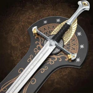 May include: A silver-toned metal sword with a black grip, featuring gold-colored accents on the pommel and crossguard. The blade is engraved with intricate patterns. It is mounted on a black, shield-shaped plaque decorated with gold filigree.