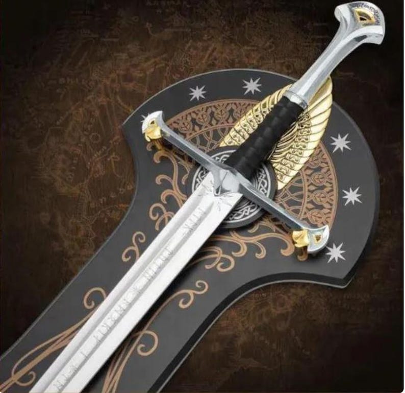 May include: A silver-toned metal sword with a black grip, featuring gold-colored accents on the pommel and crossguard. The blade is engraved with intricate patterns. It is mounted on a black, shield-shaped plaque decorated with gold filigree.