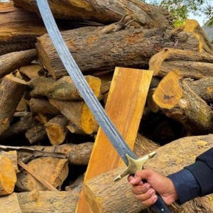 Op de afbeelding: Een gebogen zwaard met een patroonstalen lemmet, een messing pareerstang en een donker handvat. Het lemmet heeft een zichtbare textuur in Damascus-stijl over de gehele lengte, terwijl het gevest een messing pommel en pareerstang bevat.