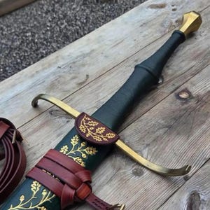 Peut inclure: Une épée avec une poignée gainée de cuir noir et un pommeau doré. La garde est en métal doré. Le fourreau vert foncé présente des motifs de feuilles de chêne et de glands dorés, maintenu par des lanières en cuir marron avec un empiècement assorti.