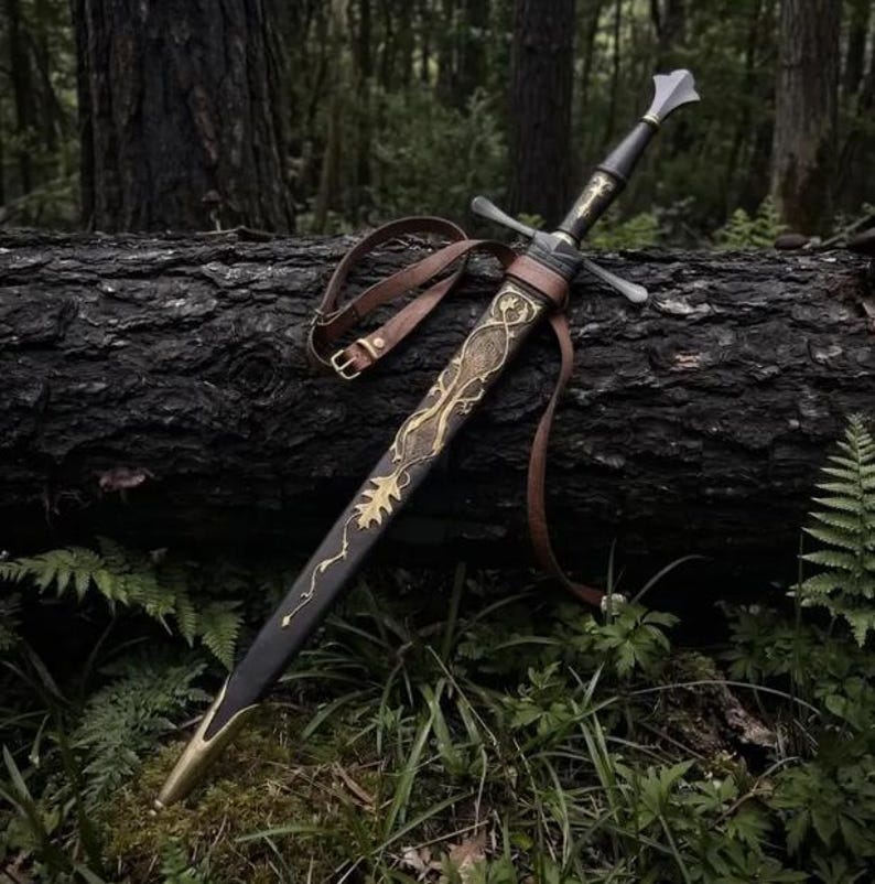 May include: A longsword with a dark handle and a silver-toned pommel, resting in a black scabbard. The scabbard features intricate gold-colored metal filigree in a vine and leaf pattern, with a matching gold-colored tip and a brown leather strap.