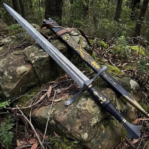 May include: A long sword with a textured, patterned steel blade, a black hilt with gold-colored accents, and a matching black scabbard featuring intricate gold-colored filigree and a brown leather strap, resting on a mossy, uneven rock surface.