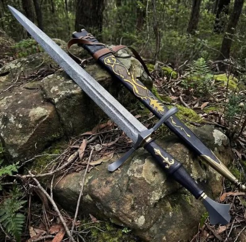 May include: A long sword with a textured, patterned steel blade, a black hilt with gold-colored accents, and a matching black scabbard featuring intricate gold-colored filigree and a brown leather strap, resting on a mossy, uneven rock surface.