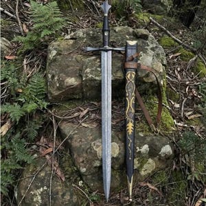 May include: A double-edged steel sword with a black grip and a crossguard, positioned next to a black scabbard featuring gold-colored filigree patterns and a brown leather strap. The sword blade has a textured, mottled finish along its length.