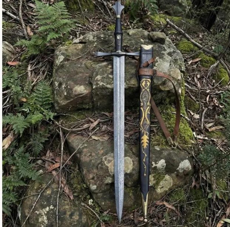 May include: A double-edged steel sword with a black grip and a crossguard, positioned next to a black scabbard featuring gold-colored filigree patterns and a brown leather strap. The sword blade has a textured, mottled finish along its length.