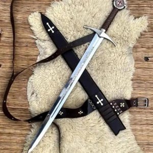 May include: A silver-toned steel sword with a brown leather-wrapped grip and a circular pommel featuring a red cross emblem. The sword is accompanied by a black leather scabbard and a matching belt, both decorated with small white cross inlays.