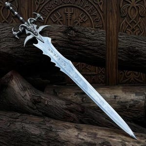 May include: A silver-toned metal sword featuring a detailed ram skull hilt with curved horns and a dark wrapped handle. The blade is long, pointed, and etched with runic-style inscriptions along the upper section near the crossguard.