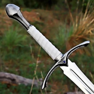 Pode incluir: Uma espada com um pomo de metal prateado polido, um punho envolto em branco e uma guarda com uma pequena incrustação circular azul no centro. A lâmina é feita de metal prateado reflexivo com um sulco central ao longo do seu comprimento.
