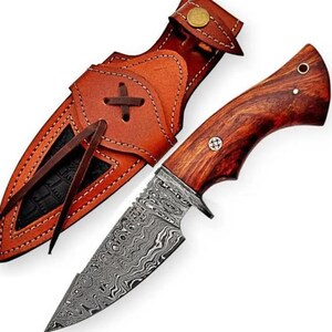 May include: A fixed-blade knife featuring a patterned Damascus steel blade and a polished reddish-brown wood handle with two metal pins. The knife is accompanied by a tan leather sheath with decorative stitching, a brass snap, and leather ties.