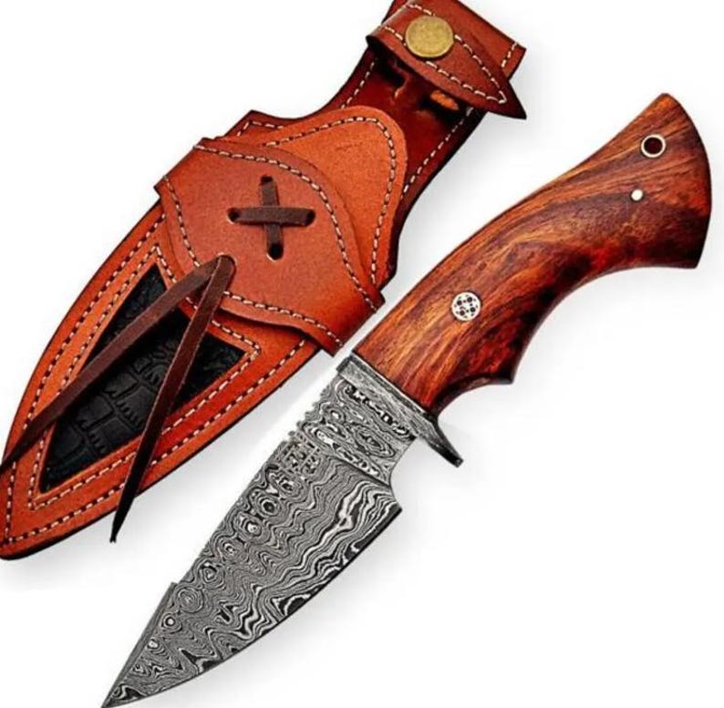 May include: A fixed-blade knife featuring a patterned Damascus steel blade and a polished reddish-brown wood handle with two metal pins. The knife is accompanied by a tan leather sheath with decorative stitching, a brass snap, and leather ties.