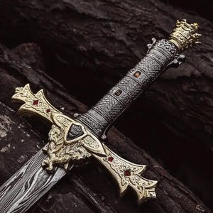 Op de afbeelding: Een decoratief zwaard met een damaststalen lemmet met een golvend patroon. Het gevest heeft een goudkleurige pareerstang met leeuwenmotieven en rode accenten, een textuurrijke donkere metalen greep met kleine steentjes en een gouden kroonknop.