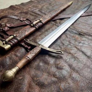 Könnte beinhalten: Ein Stahlschwert mit zweischneidiger Klinge, Messing-Parierstange und Holzgriff, der in einem Messingknauf endet. Das Schwert wird von einer braunen Lederscheide mit geprägten Mustern, Messingakzenten und Lederriemen zum Tragen begleitet.