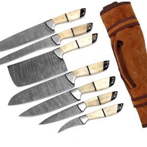 Può includere: Un set di sei coltelli da cucina con lame in acciaio damasco a motivi e manici color crema con accenti scuri, accompagnato da una custodia arrotolabile in pelle marrone con manico e chiusure a cinghia per organizzare il set di coltelli.