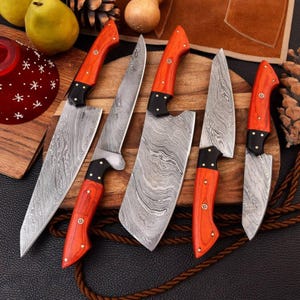 Könnte beinhalten: Ein Set aus fünf Küchenmessern mit Damaststahlklingen mit Wellenmuster und leuchtend orangefarbenen Holzgriffen, die mit Messingnieten befestigt sind. Die Messer variieren in Klingenform und Größe, vom großen Kochmesser bis zum kleinen Schälmesser.