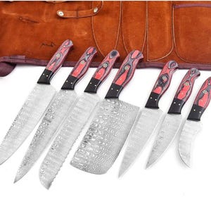 Può includere: Un set di sette coltelli da cucina con lame in acciaio damasco dal motivo ondulato e manici in legno rosso e nero con rivetti in ottone. I coltelli sono disposti in fila, tra cui una mannaia, coltelli da chef e spelucchini, con un rotolo in pelle.